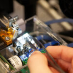 Image shows a close up of a chip being put on a probe station, Scott is holding tweezers which have the chip between them