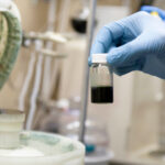 Delwin's hand in a latex glove holds OEO materials in a small glass vial near a machine under the fume hood in NLM's wet lab