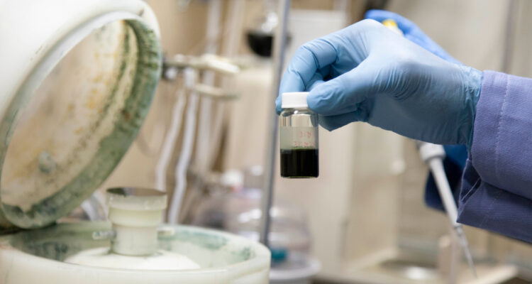 Delwin's hand in a latex glove holds OEO materials in a small glass vial near a machine under the fume hood in NLM's wet lab